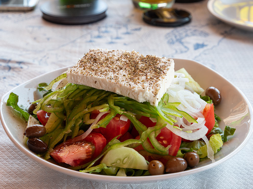 Mixture of tomatoes, cucumber, green peppers, lettuce and olives, with a slice of feta seasoned with oregano in a white bowl.