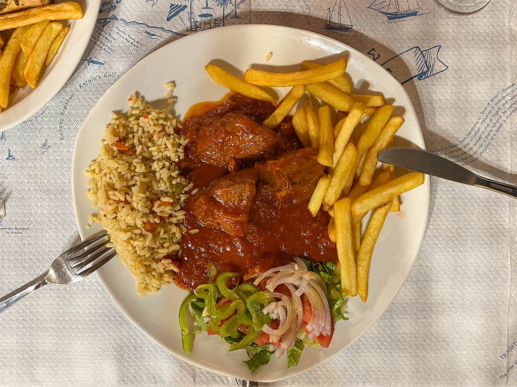Large chunks of meat in a deep red tomato sauce served with rice, fries and mixed salad on a white plate.