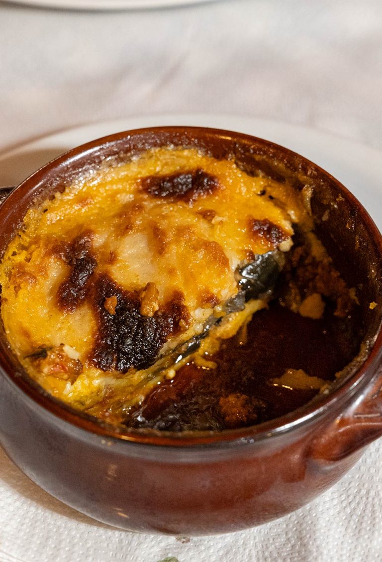 Meat and vegetable meal with a golden béchamel crust in a round brown glazed clay pot, showing the layered construction.