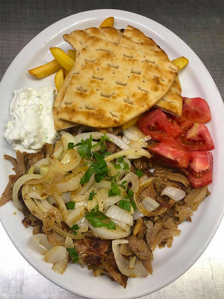 Gyros plate of cooked sliced onions and meat, sliced red tomatoes, spoonful of creamy tzatziki, fries beneath pieces of pita bread.