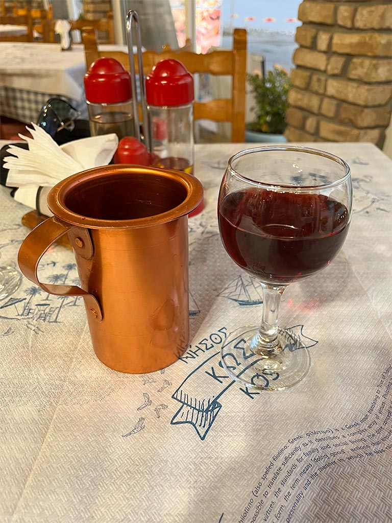 Red wine in a glass and copper colour serving jug, on a white paper tablecloth with Kos in blue print.