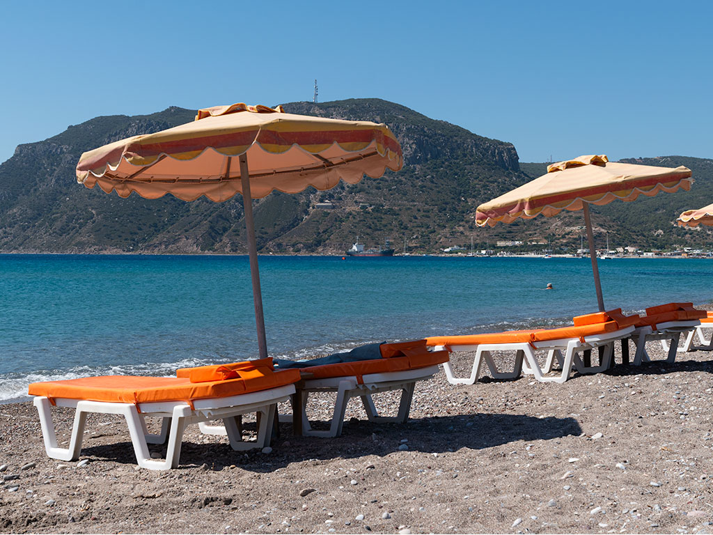 Rainbow Taverna sunbeds for hire with cushions and parasols on the beach overlooking the sea and a cloudless blue sky.