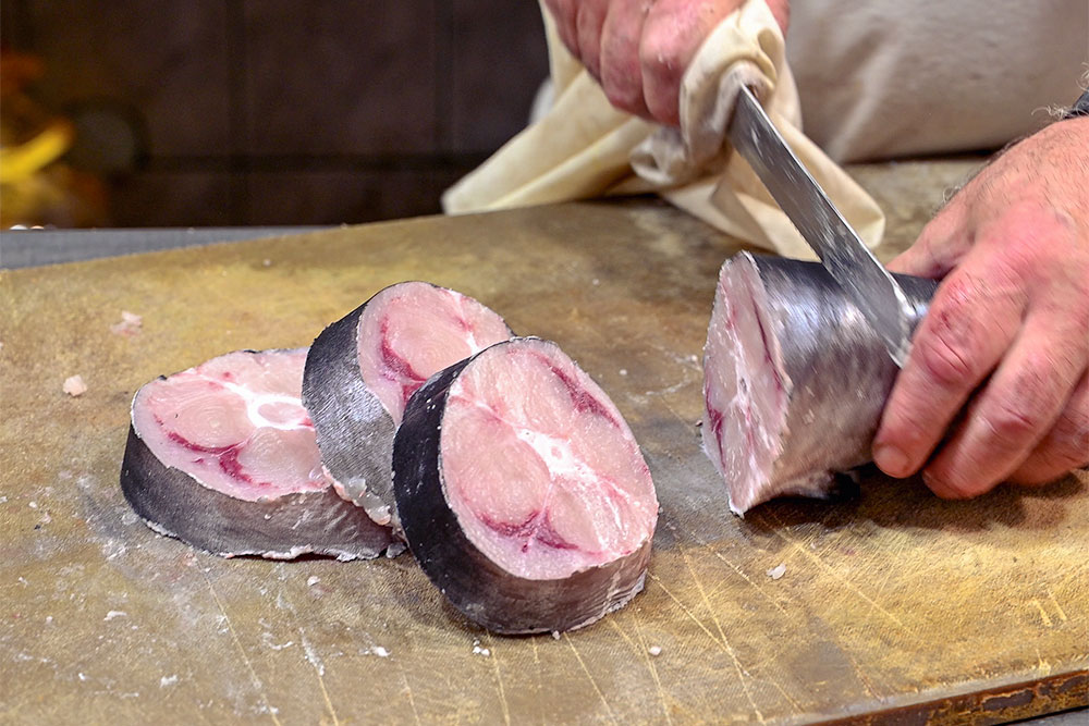 Three medallions of Swordfish on a wooden chopping board. The chef’s hands with knife ready to prepare the next one.