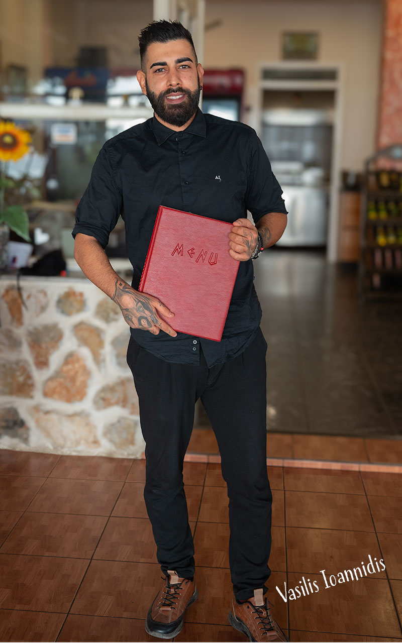 Standing waiter in black uniform with maroon coloured menu holder smiling and posing for the camera.